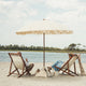 Two people sitting under a large luxury beach umbrella on a sandy beach with water and trees in the background.
