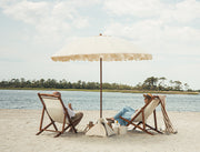 Two people sitting under a large luxury beach umbrella on a sandy beach with water and trees in the background.