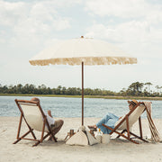 Two people sitting under a large luxury beach umbrella on a sandy beach with water and trees in the background.