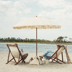 Two people sitting under a large luxury beach umbrella on a sandy beach with water and trees in the background.