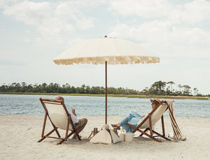 Two people sitting under a large luxury beach umbrella on a sandy beach with water and trees in the background.
