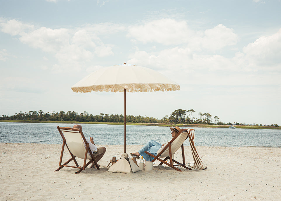 Two people sitting under a large luxury beach umbrella on a sandy beach with water and trees in the background.