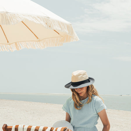 Woman in a light blue dress and hat sitting under a large luxury beige beach umbrella on a sandy beach.